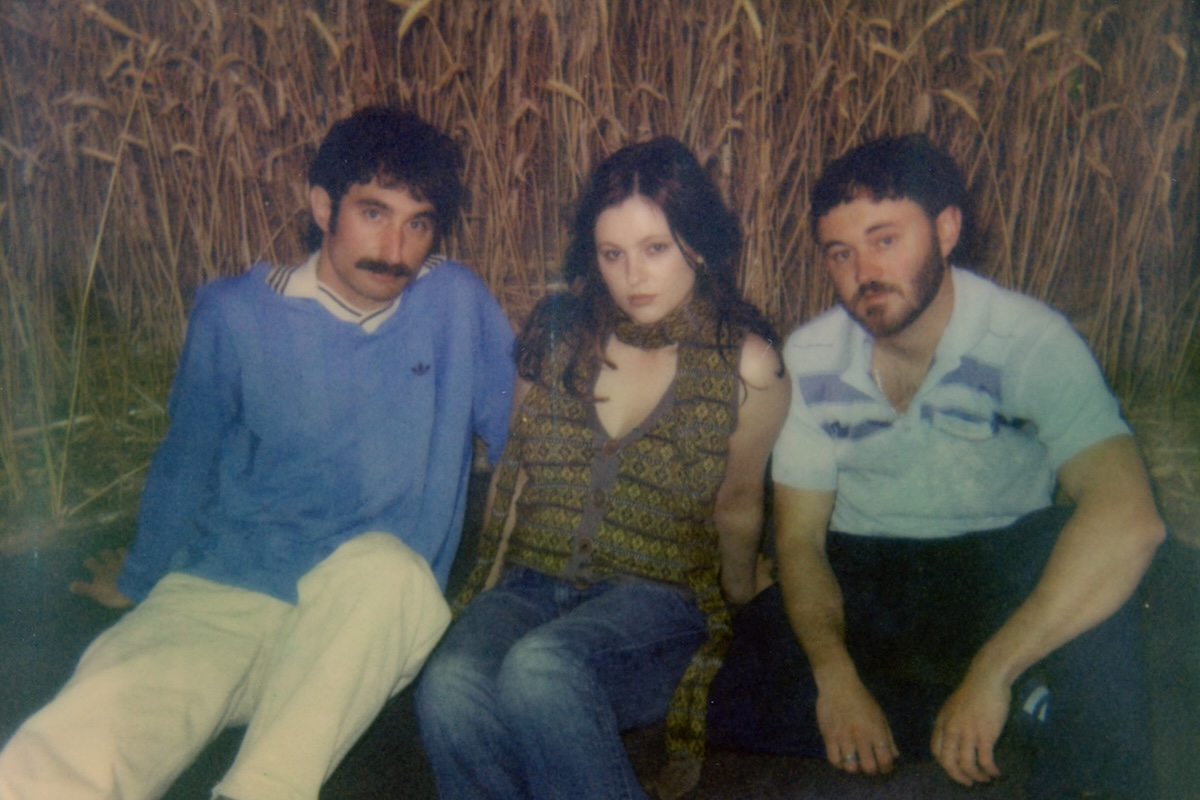 Polaroid photo of Australian indie pop band San Cisco sitting together outdoors in front of tall grass in a casual group portrait.